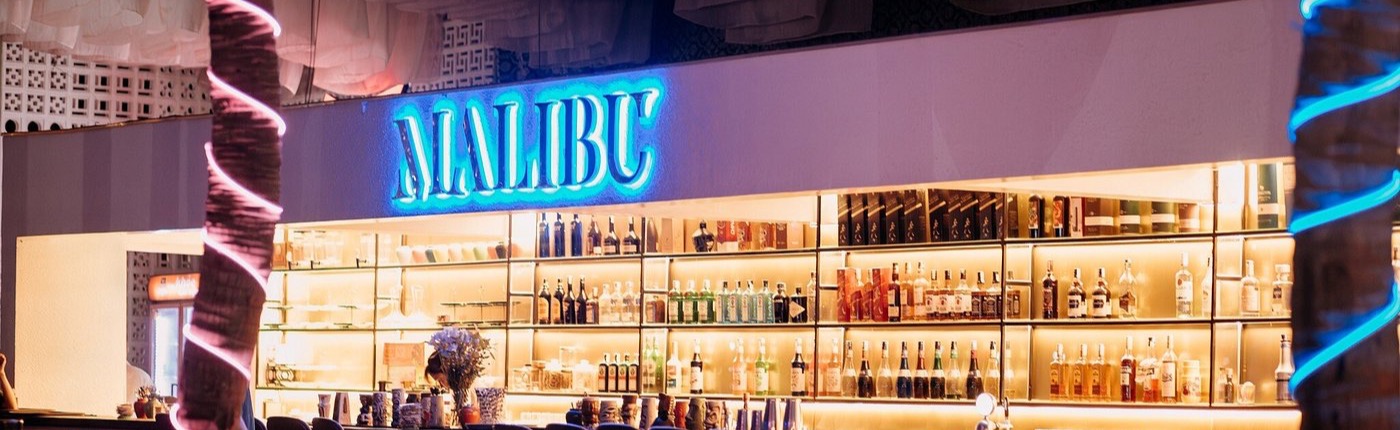 Malibu Beach Club, Đà Nẵng - quầy bar với biển hiệu neon MALIBU và hàng kệ rượu được chiếu sáng ấm áp, hai cây dừa hai bên